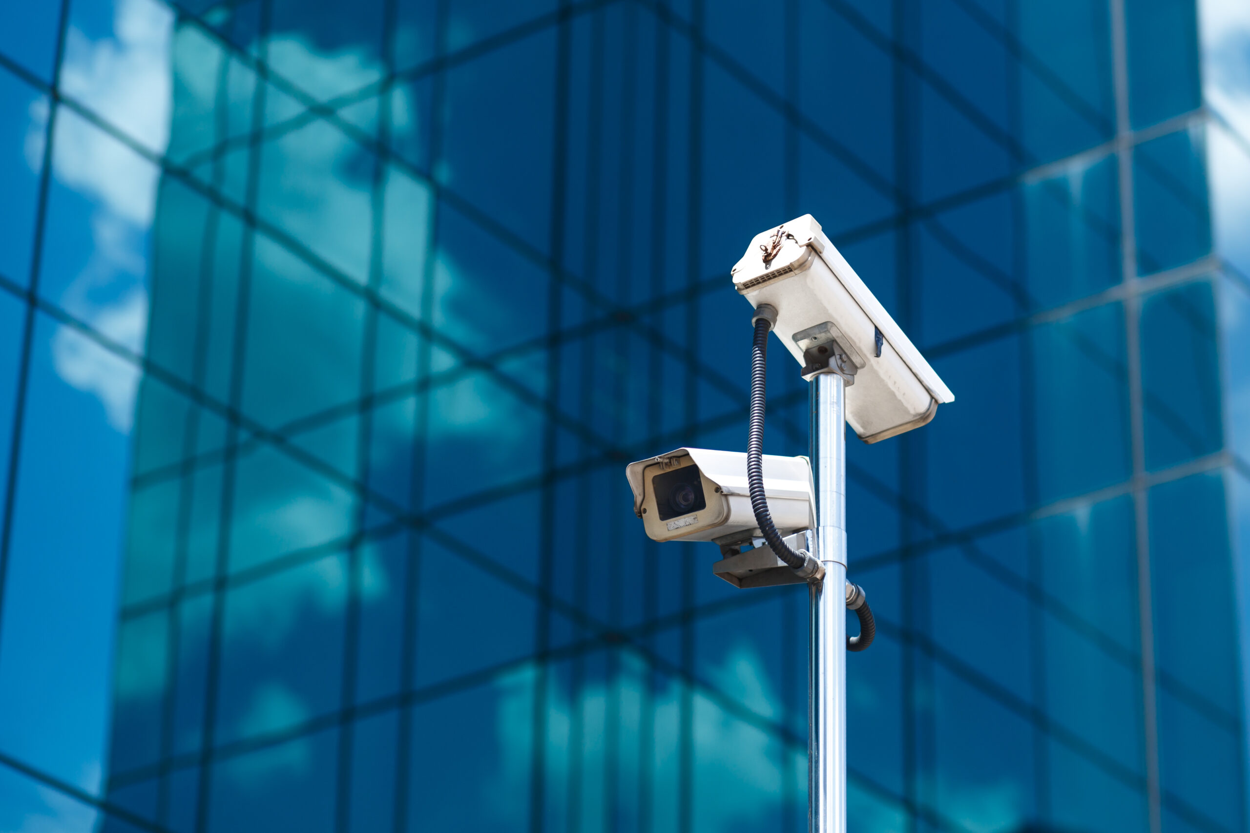Poteau avec deux caméras de vidéosurveillance blanches devant un grand immeuble de bureaux en verre.